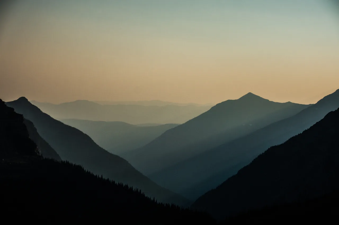 Layered Glacier mountain ridgelines fading into smoky dusk, warm sky over teal shadowed peaks