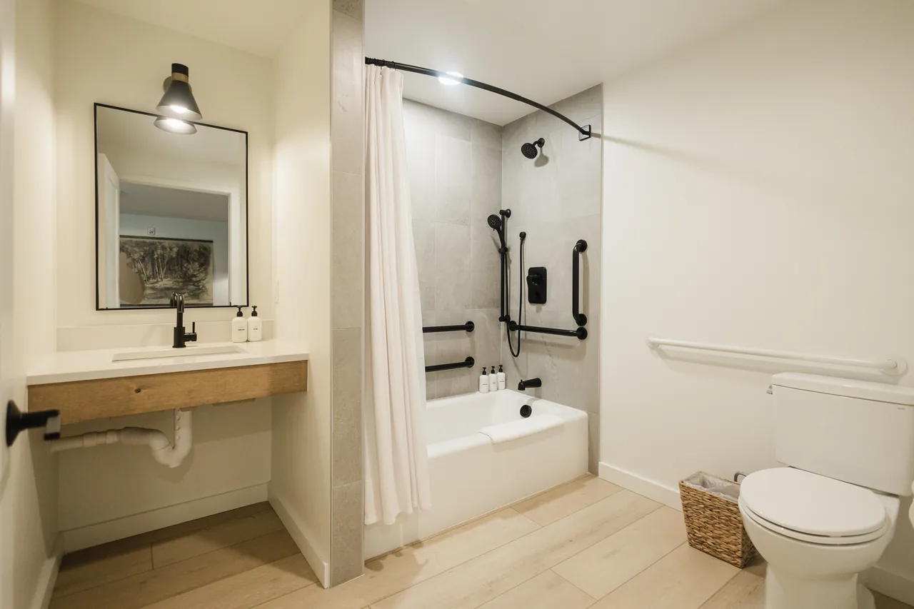 Accessible bathroom with floating vanity, warm wall sconce, tub-shower with black fixtures and grab bars