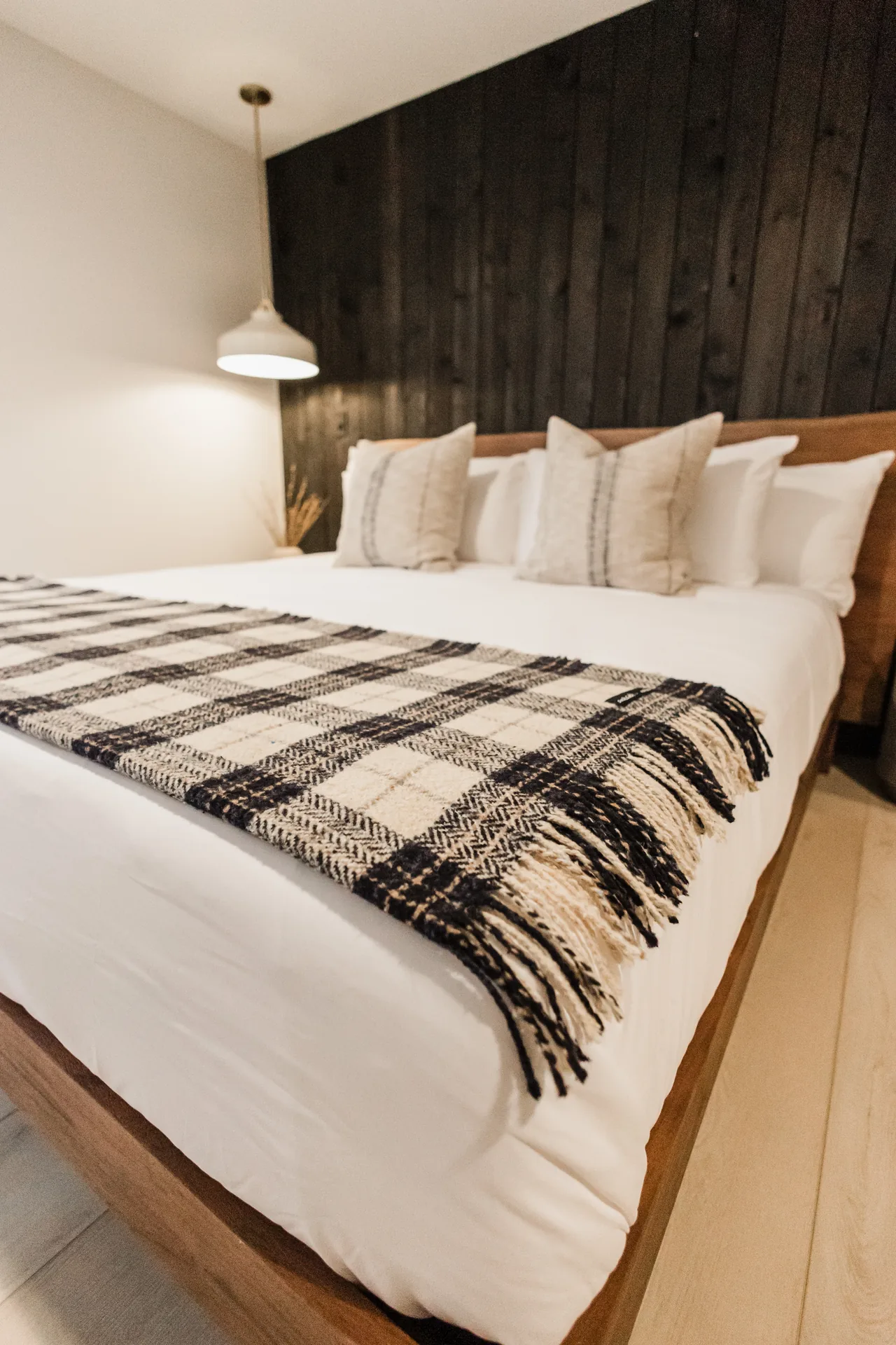 King bed with plaid fringe throw over white linens, walnut headboard against dark shiplap, glowing pendant