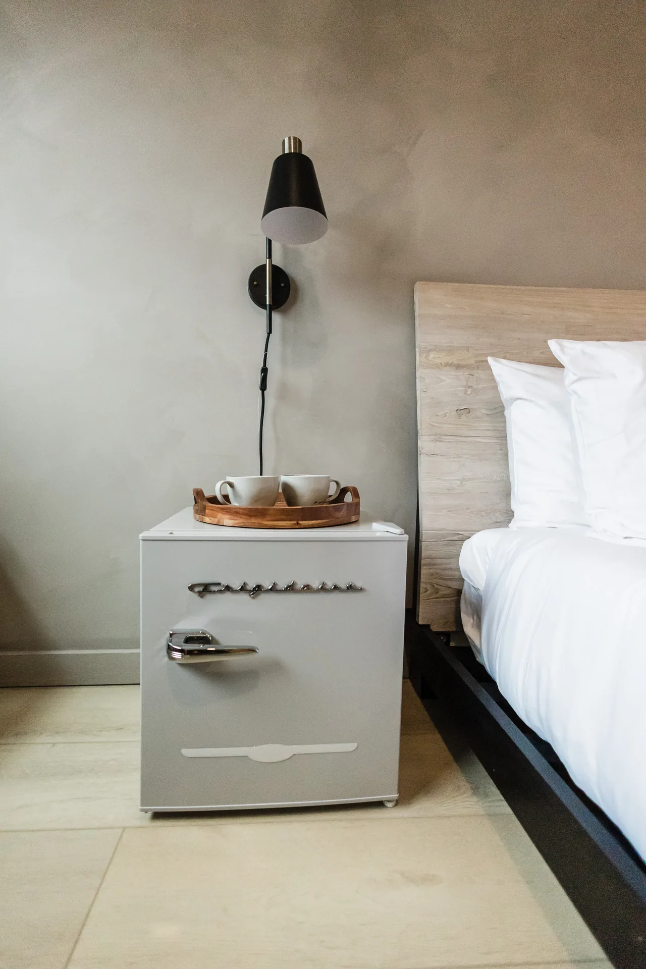 Retro-style mini fridge nightstand topped with a wood tray and ceramic mugs beside the king bed