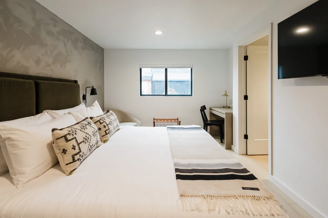 Low-angle view of king bed with layered white pillows, woven throw blanket, and open bathroom beyond