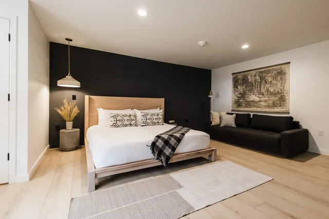 King bed with woven throw pillows against charcoal shiplap wall, pendant light, and dark sofa alongside large-format art