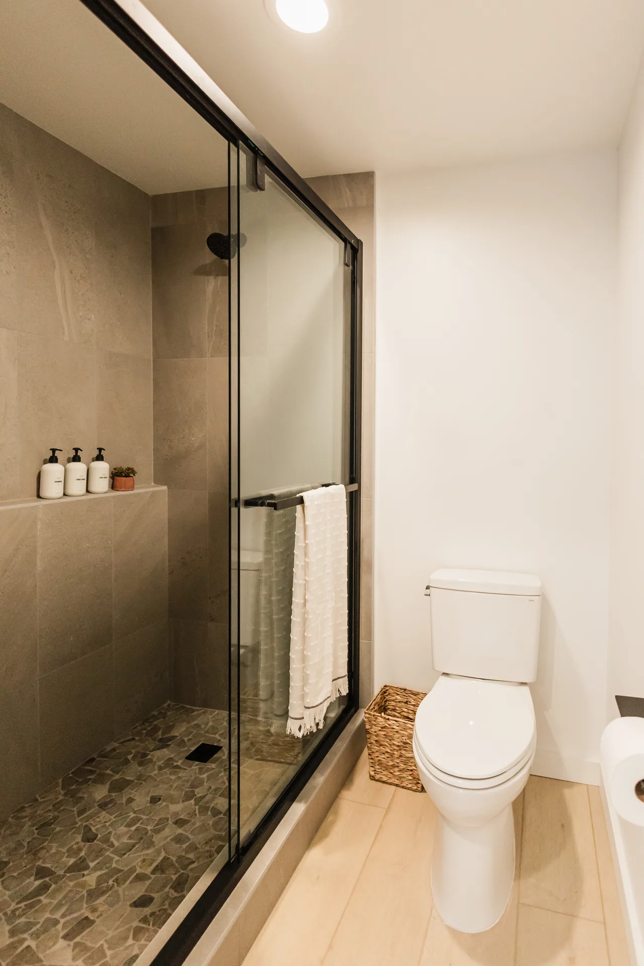 Walk-in shower with large-format taupe tile, pebble stone floor, glass enclosure, and branded amenities on built-in shelf