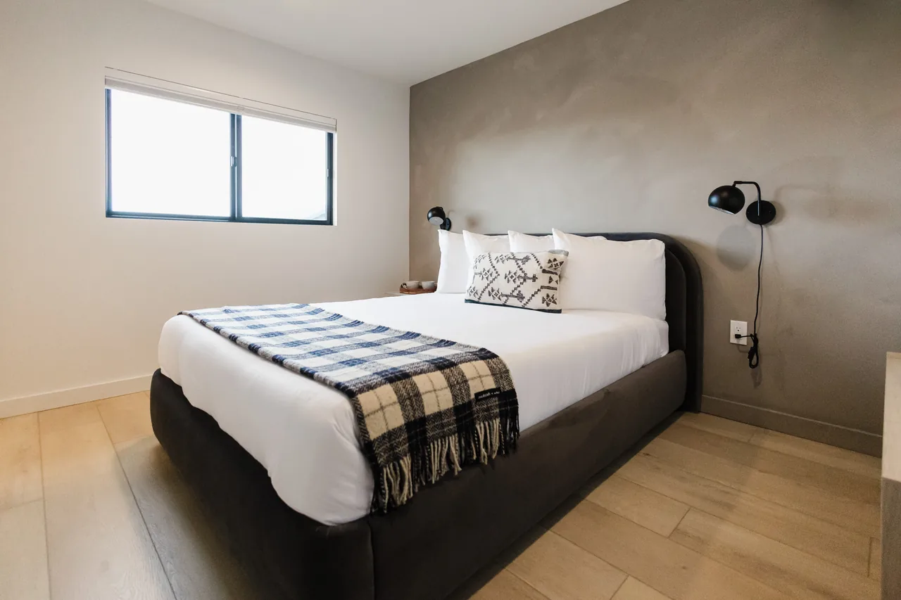 Queen bed with blue plaid throw against grey plaster accent wall, black globe sconces, and light wood flooring