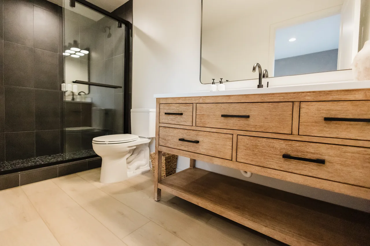 Bathroom with oak vanity, matte-black hardware, frameless glass shower with dark charcoal tile and river-stone floor