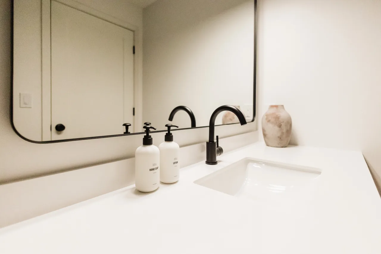 White quartz vanity counter with matte-black faucet, branded hand soap and lotion dispensers, and ceramic vase