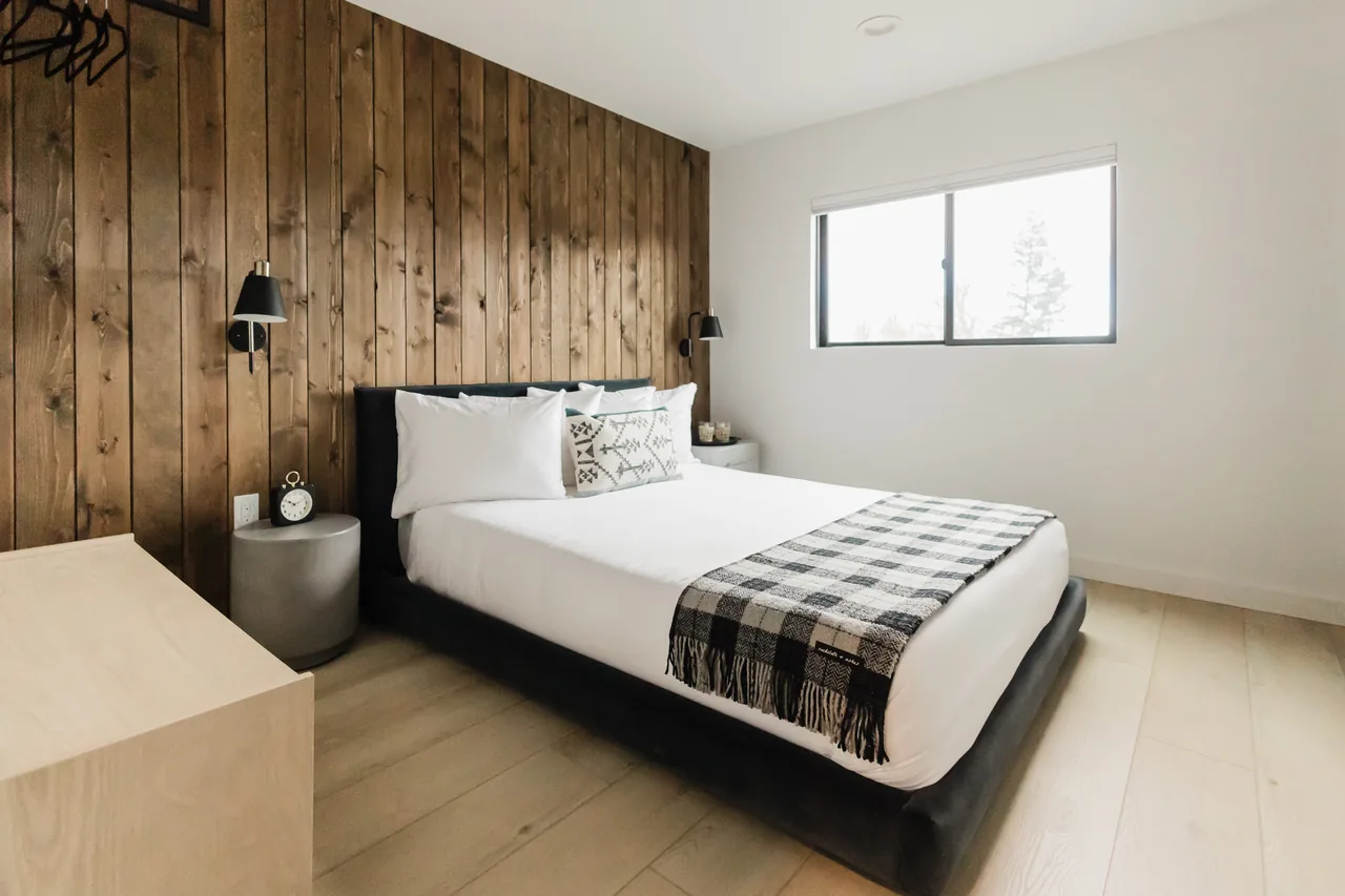 Queen bed against reclaimed wood plank wall with tartan throw blanket and black sconce lamps, light hardwood floor