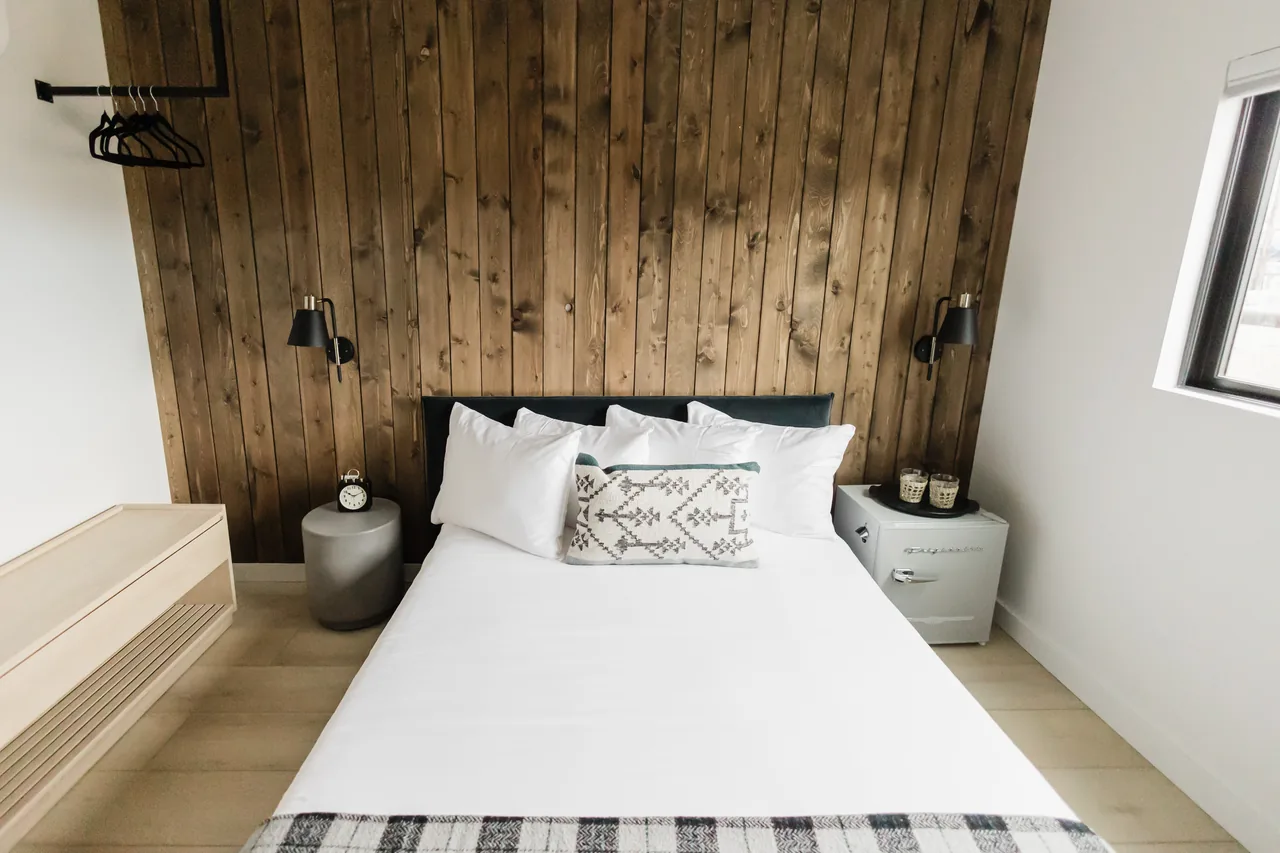 Overhead view of queen bed with white linens, tribal accent pillow, and grey Frigidaire mini-fridge nightstand against wood wall