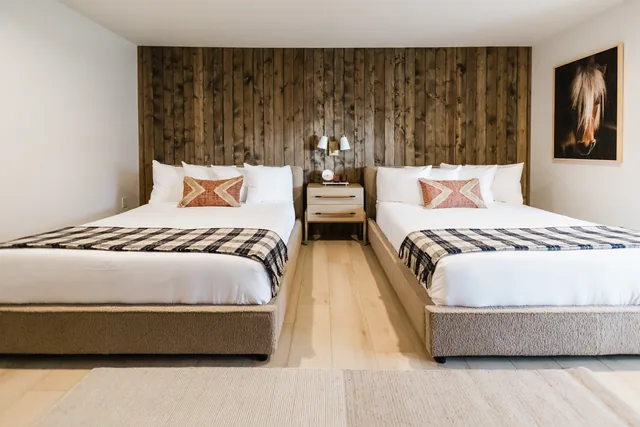 Two queen beds with plaid throws face a full-wall reclaimed wood headboard, flanked by warm brass sconces