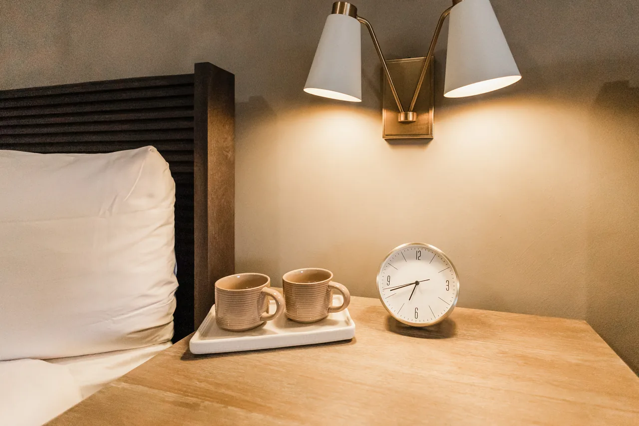 Bedside nightstand with paired ceramic mugs, brass double sconce glowing warm against a textured plaster wall