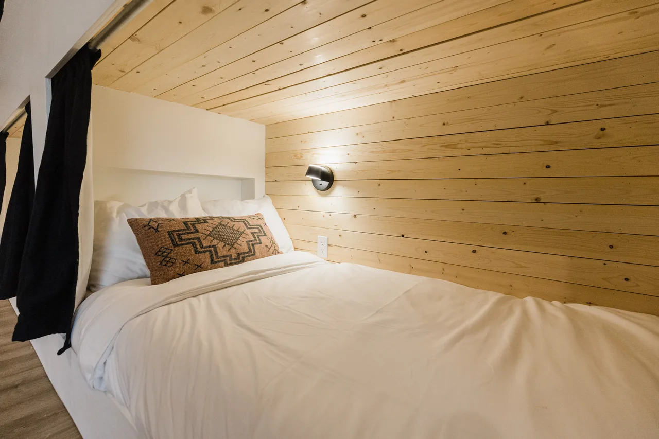 Lower bunk sleeping pod with crisp white bedding, Southwestern accent pillow, and pine shiplap ceiling with reading sconce