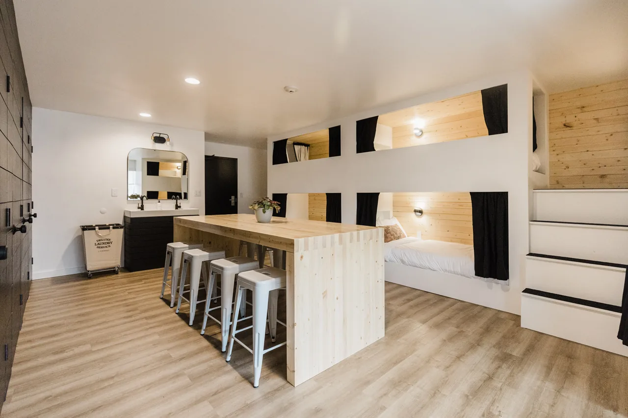 Bright bunkhouse room with built-in pine pod bunks, communal wood bar table with metal stools, and double vanity beyond