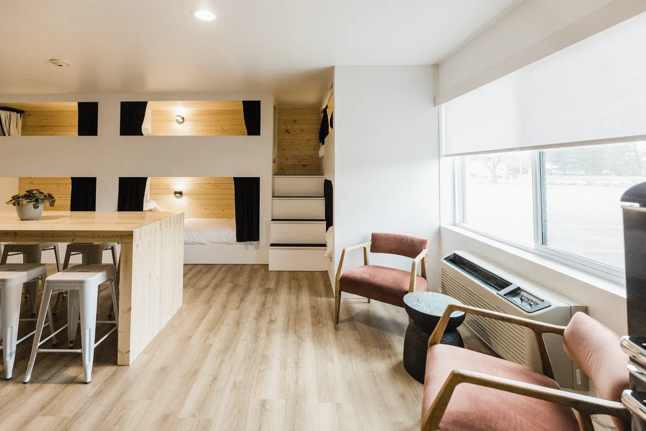 Bunkhouse seating area with warm terracotta lounge chairs by large window alongside pine bunk pods and communal table