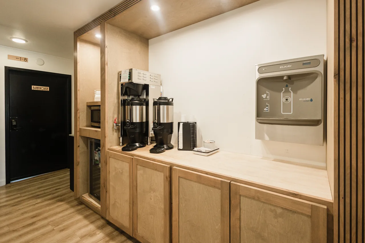Bunkhouse coffee and hydration station with warm plywood millwork, dual coffee urns, and filtered water dispenser