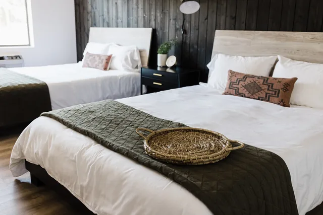 Close view of two queen beds with white linens, olive green quilts, southwestern throw pillows, and a woven tray detail