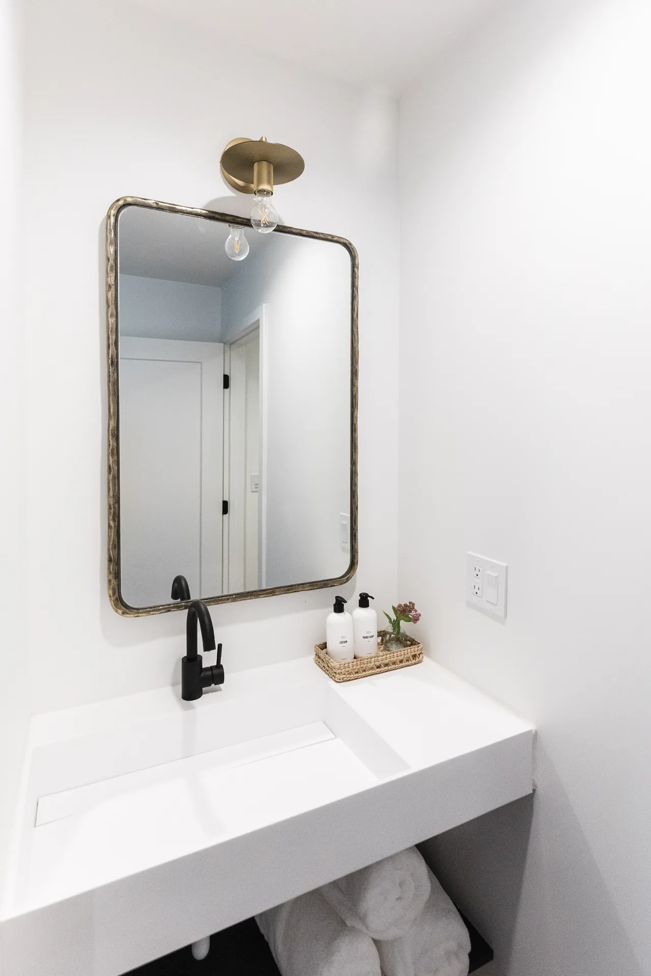 Floating white vanity with matte black faucet, aged brass mirror sconce, and neatly arranged bath amenities in a woven tray