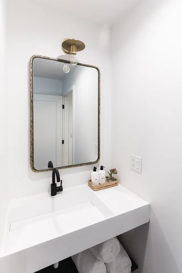 Floating white vanity with matte black faucet, aged brass mirror sconce, and neatly arranged bath amenities in a woven tray