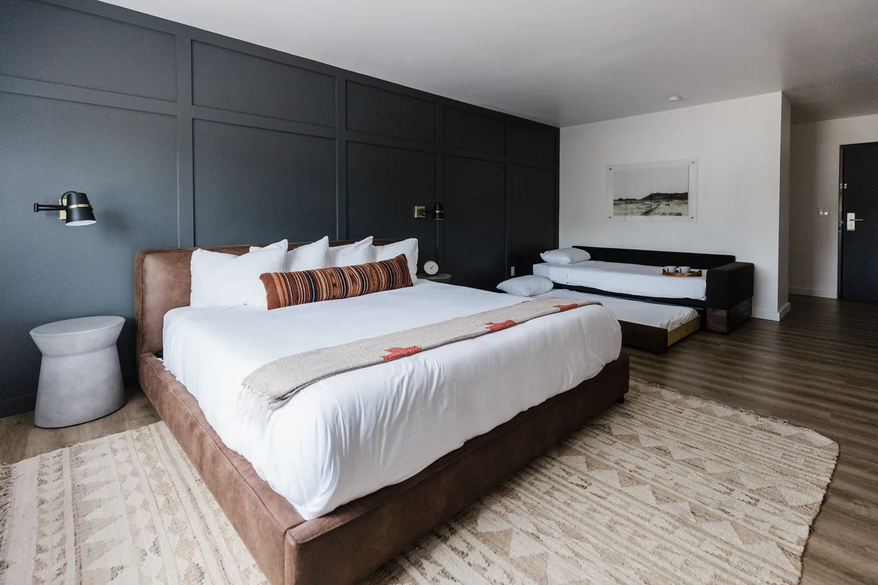 King bed with leather frame against dark paneled accent wall, trundle daybed in background, Southwestern rug
