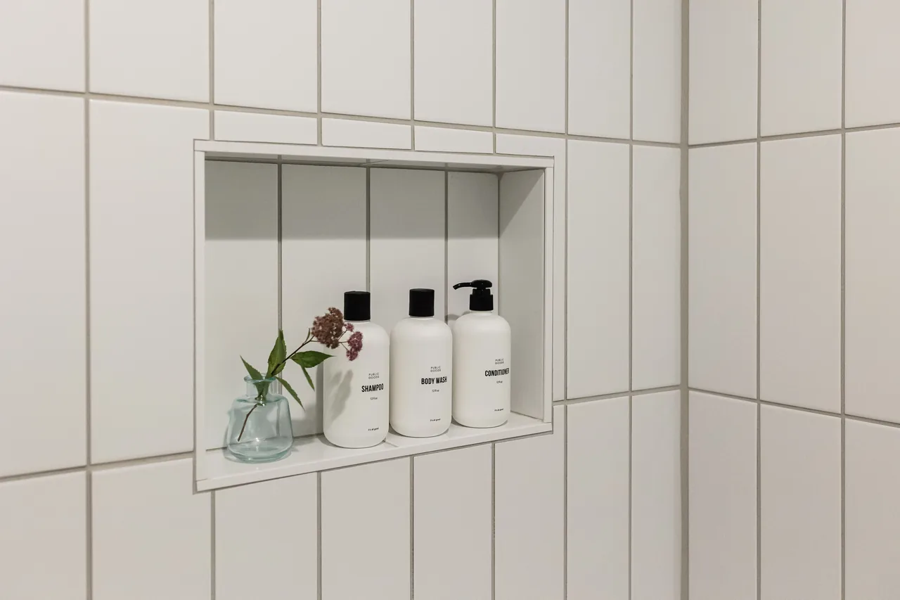 Built-in shower niche with branded shampoo, body wash, and conditioner bottles beside a small wildflower vase on white subway tile