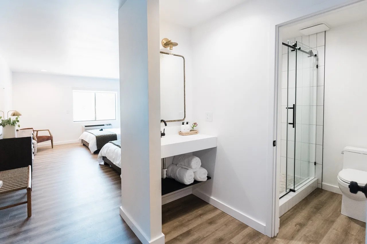 Open-plan layout showing floating vanity, glass shower, rolled towels, and king bedroom beyond in one frame