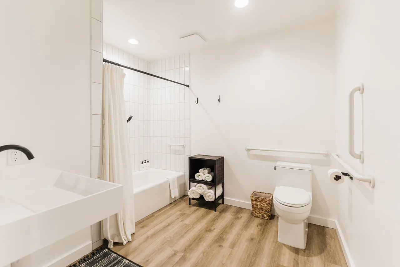 Bright ADA-accessible bathroom with white subway tile, roll-in tub, grab bars, floating vanity, and warm wood floors