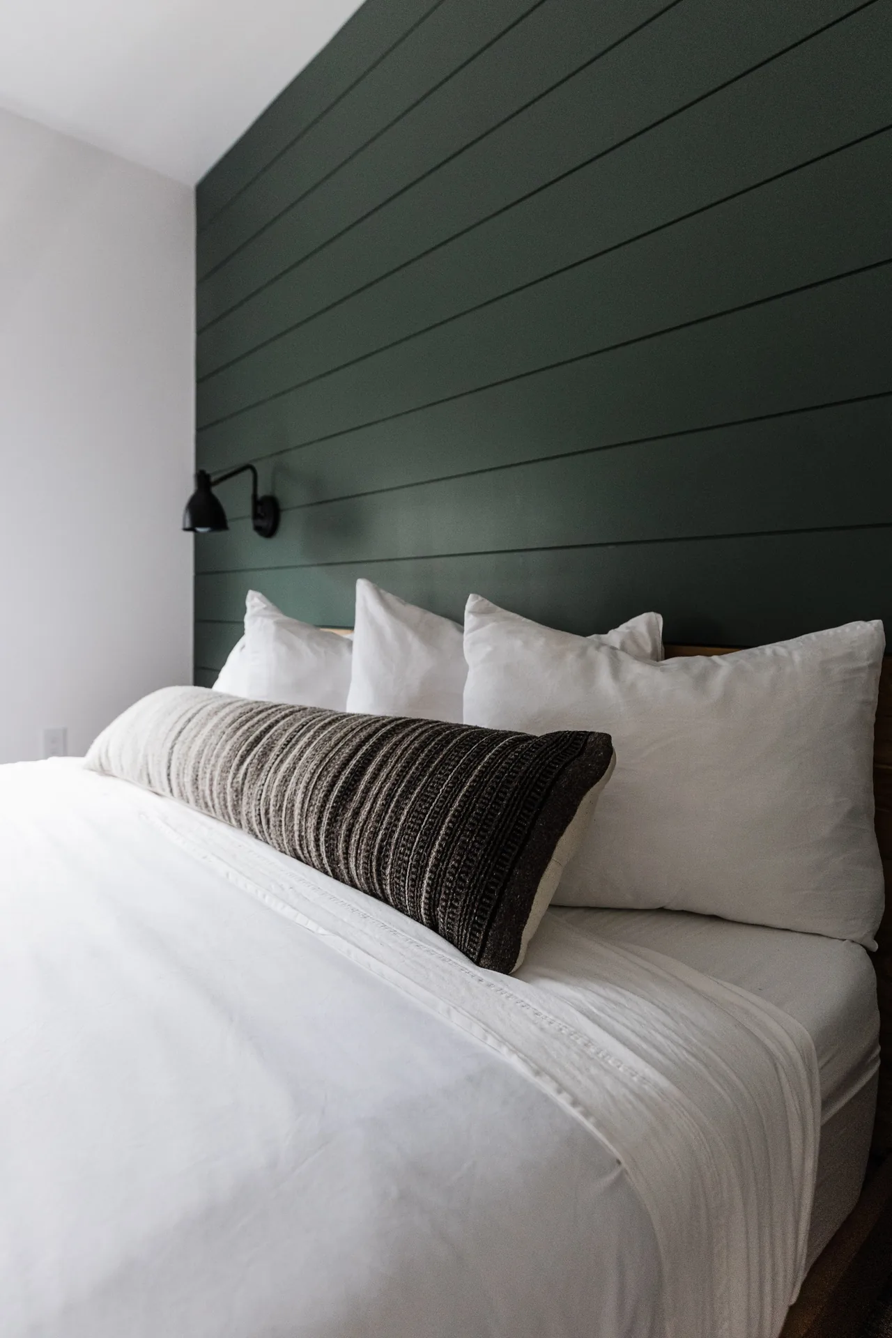 Close-up of queen bed with crisp white linens, woven lumbar pillow, and forest-green shiplap headboard wall