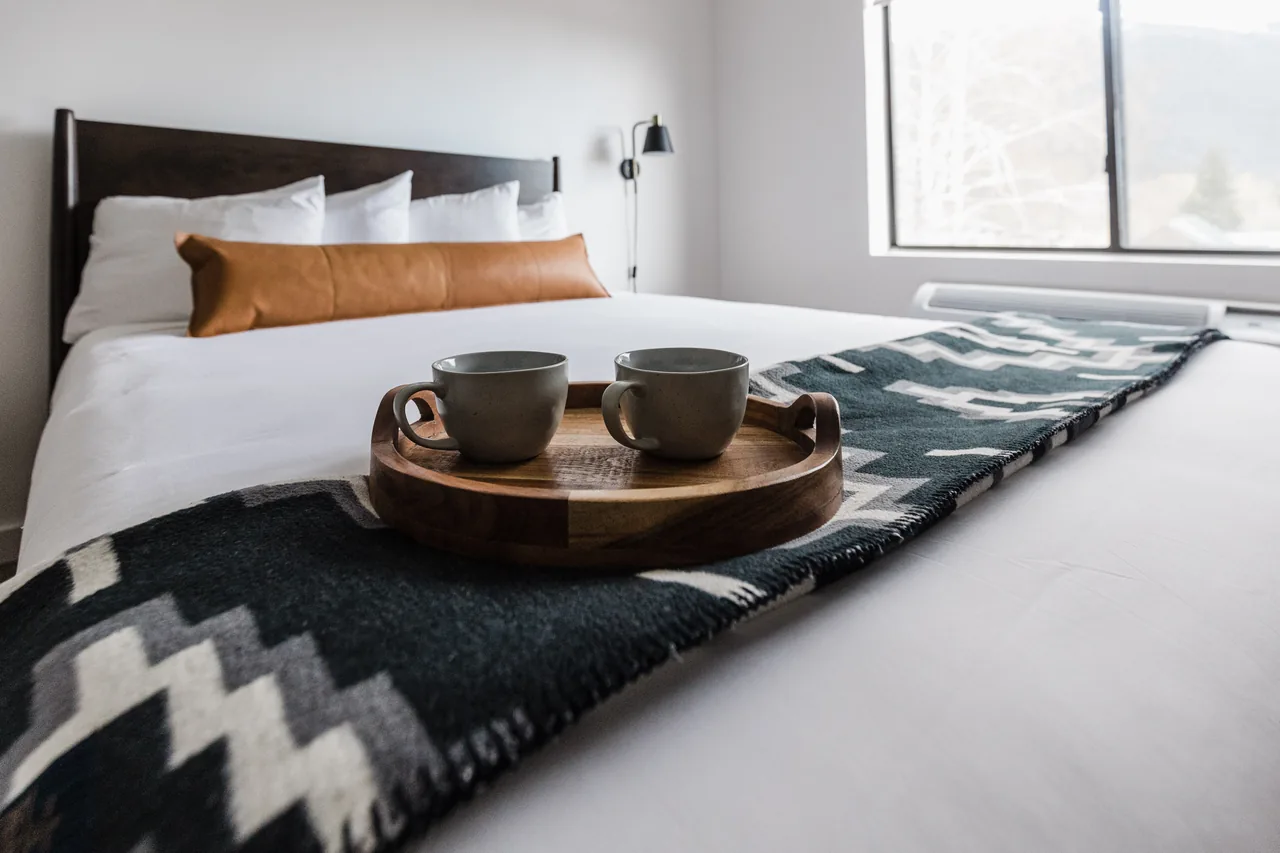 Close-up of queen bed with wooden tray, two ceramic mugs, geometric throw blanket, and mountain view window