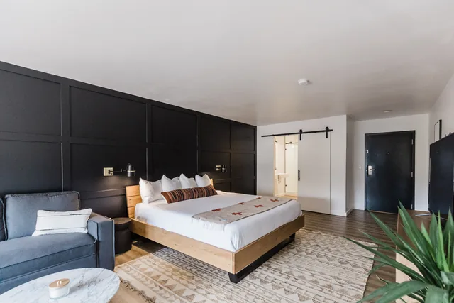 King suite with dramatic black panel accent wall, wood-platform bed, sitting area, and barn-door bath beyond